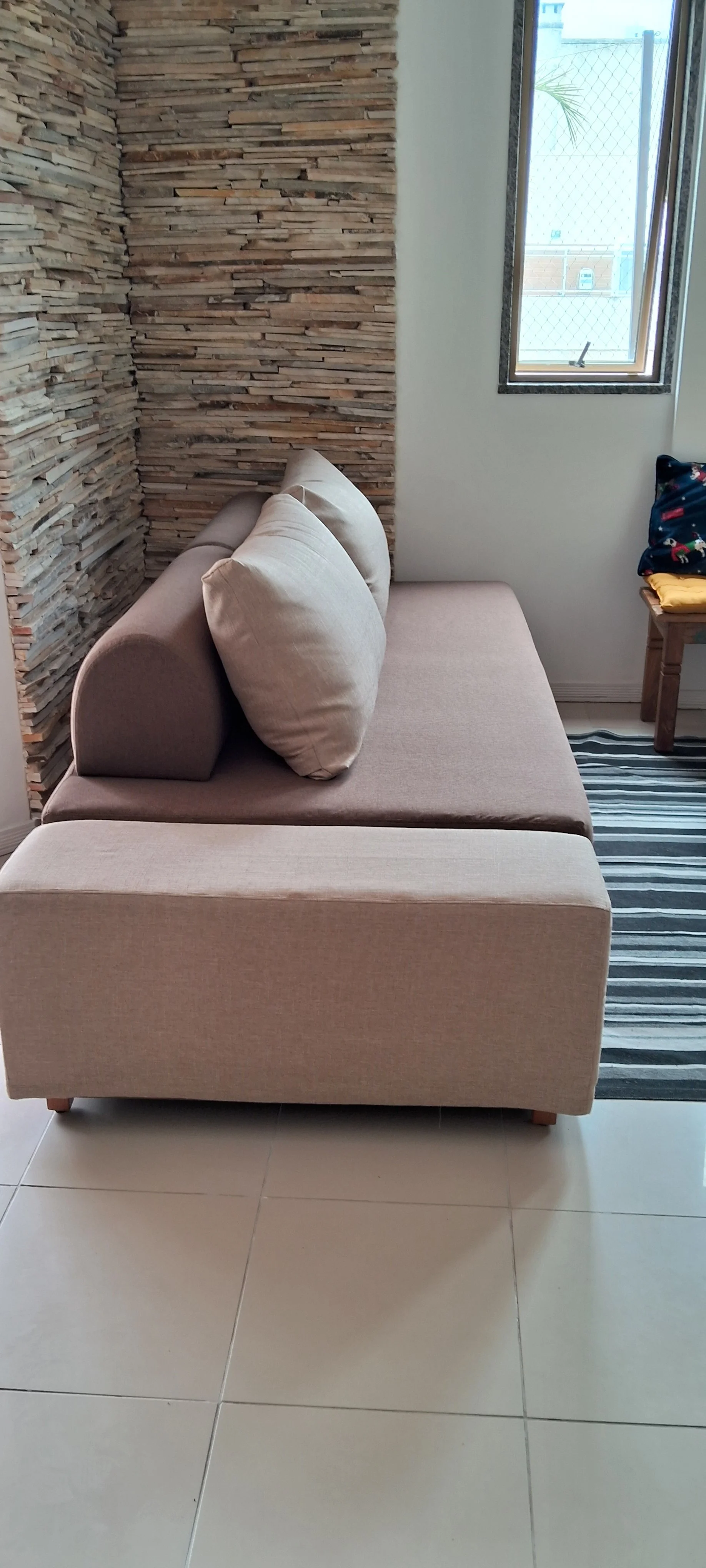 Sofá bege com almofadas na sala ao lado de uma parede com revestimento de pedras e uma janela que deixa passar luz natural.