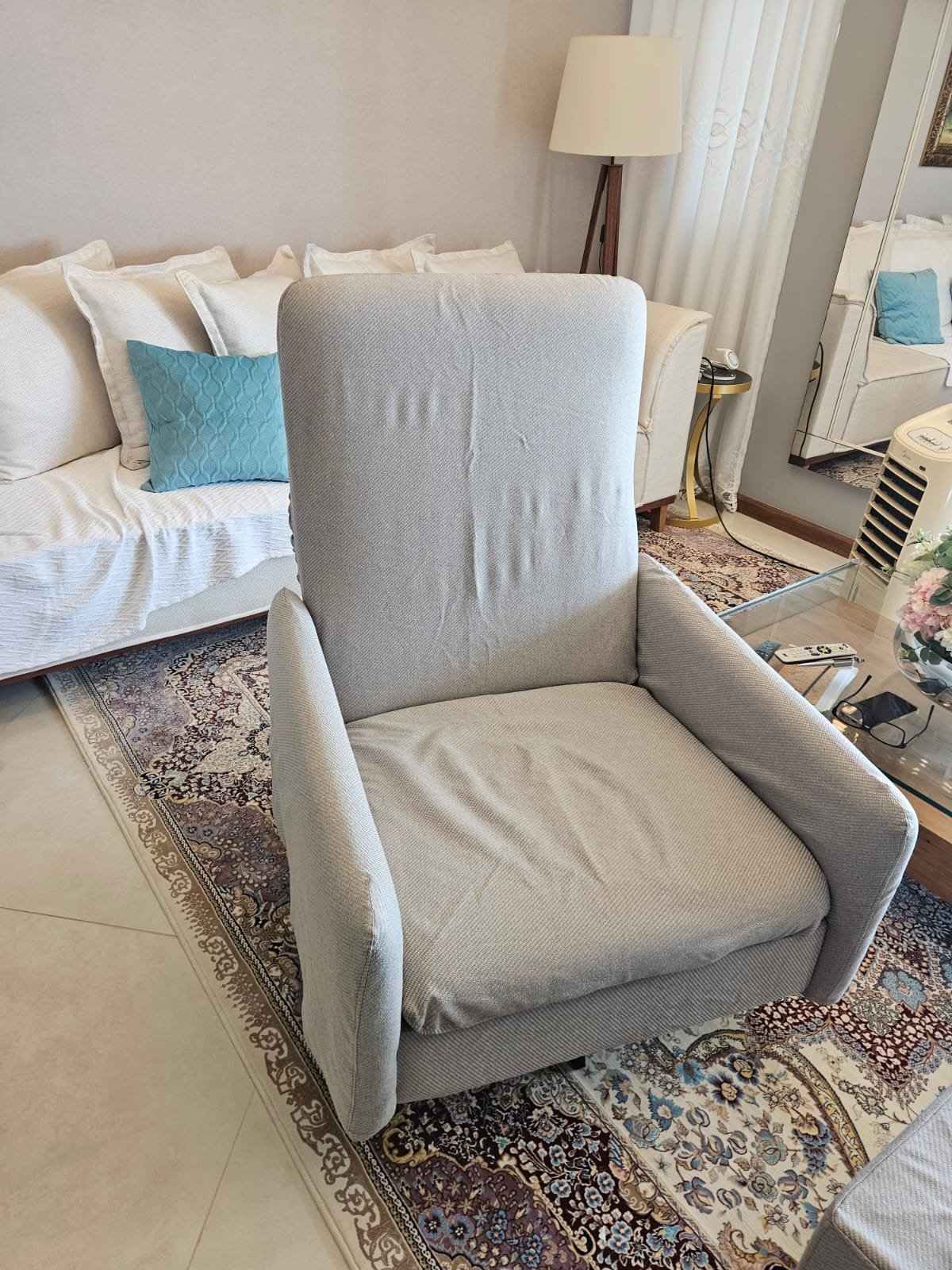 Poltrona cinza em um ambiente de sala de estar com sofá branco, tapete com motivos e uma luminária de pé ao fundo.