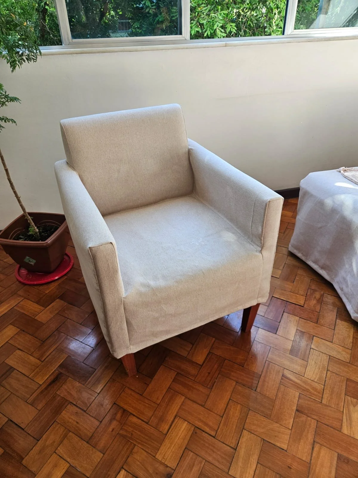 Poltrona de tecido bege ao lado de uma planta e uma mesa coberta com tecido branco, com janela ao fundo em ambiente interno com piso de madeira parquet.
