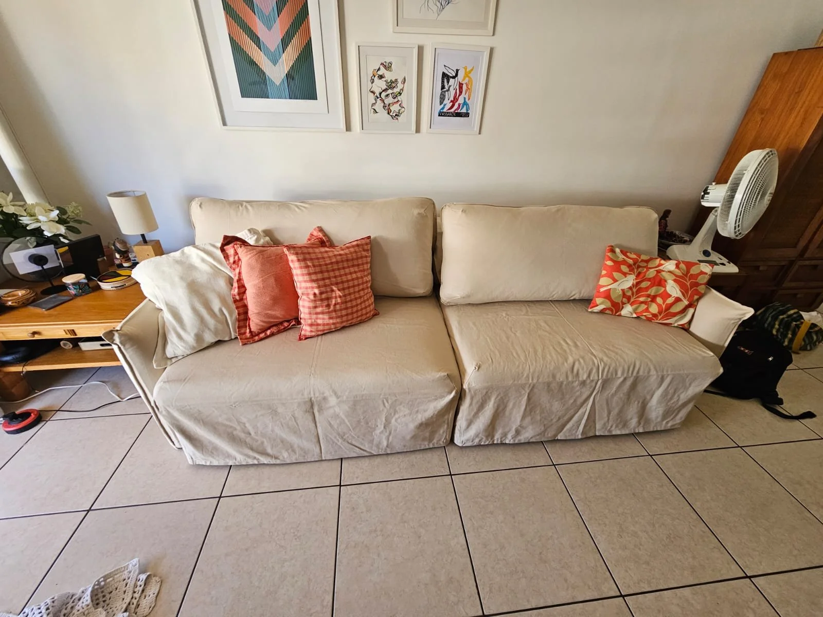 Sofá com capa bege, decorado com quatro almofadas, duas em tom de vermelho e duas em tom de creme, localizado em sala de estar. Ao lado, uma mesa de madeira com objetos diversos e uma planta com flores brancas. No fundo, quadros decorativos na parede