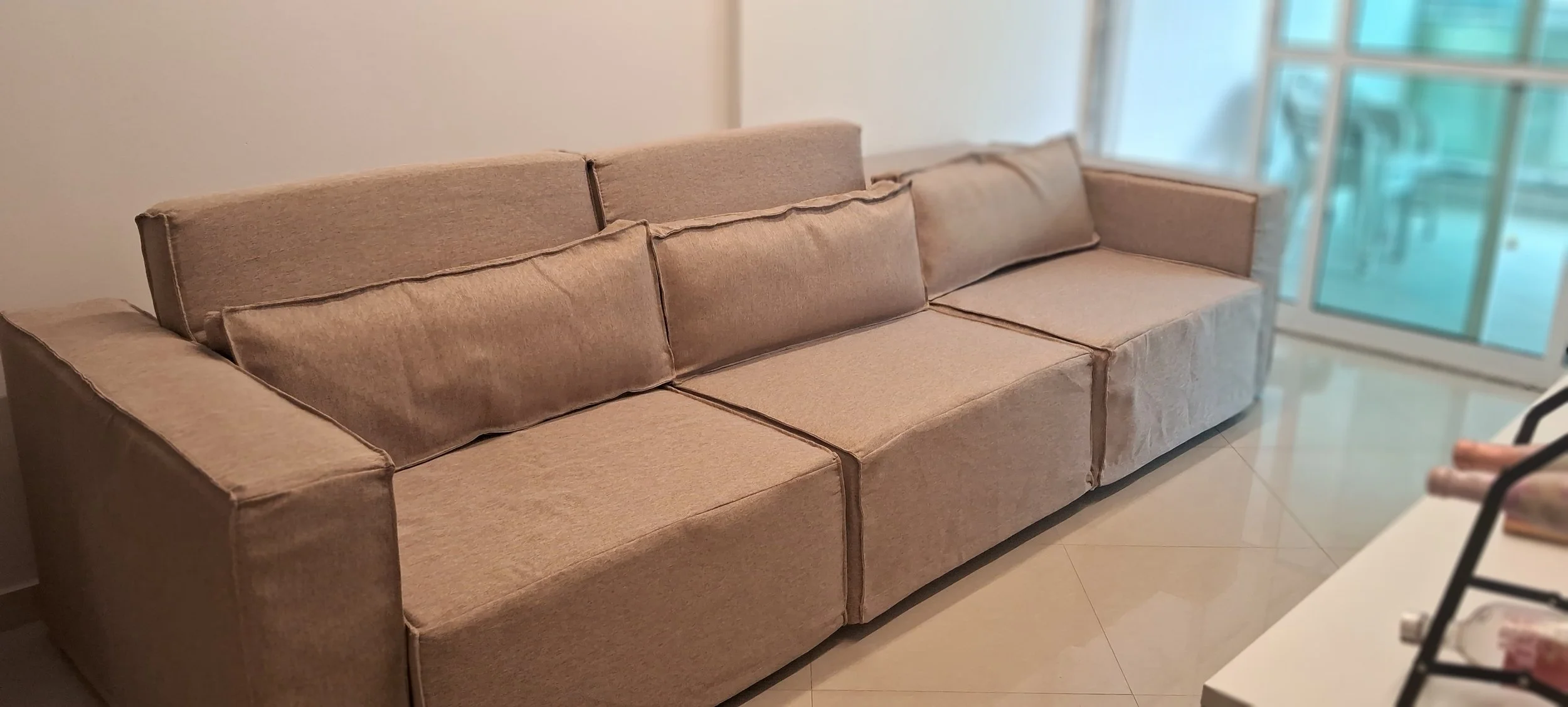 Sofá grande de tecido bege na sala, com almofadas, perto de janelas de vidro com cortinas transparentes.