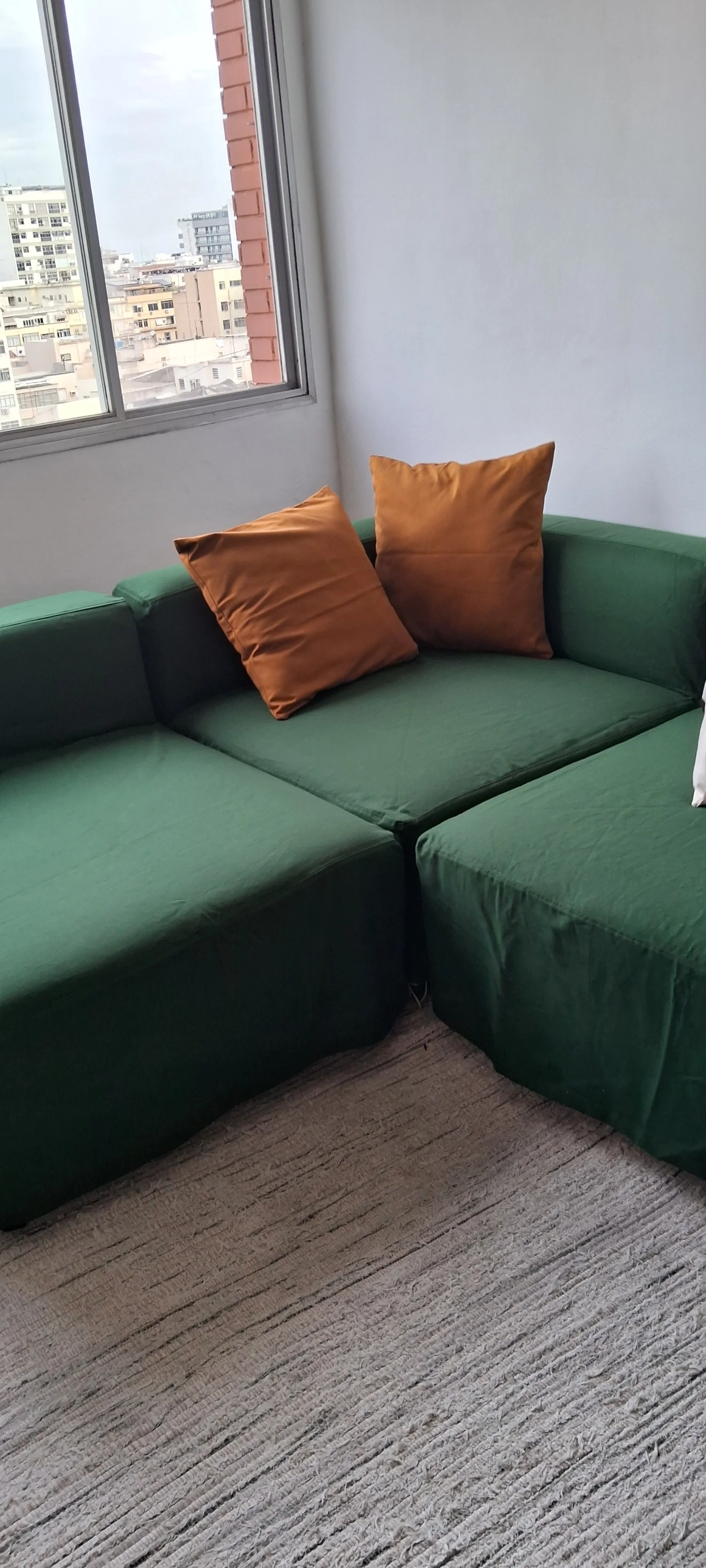 Sofá verde com dois travesseiros marrons em uma sala perto de uma janela com vista para edifícios urbanos.