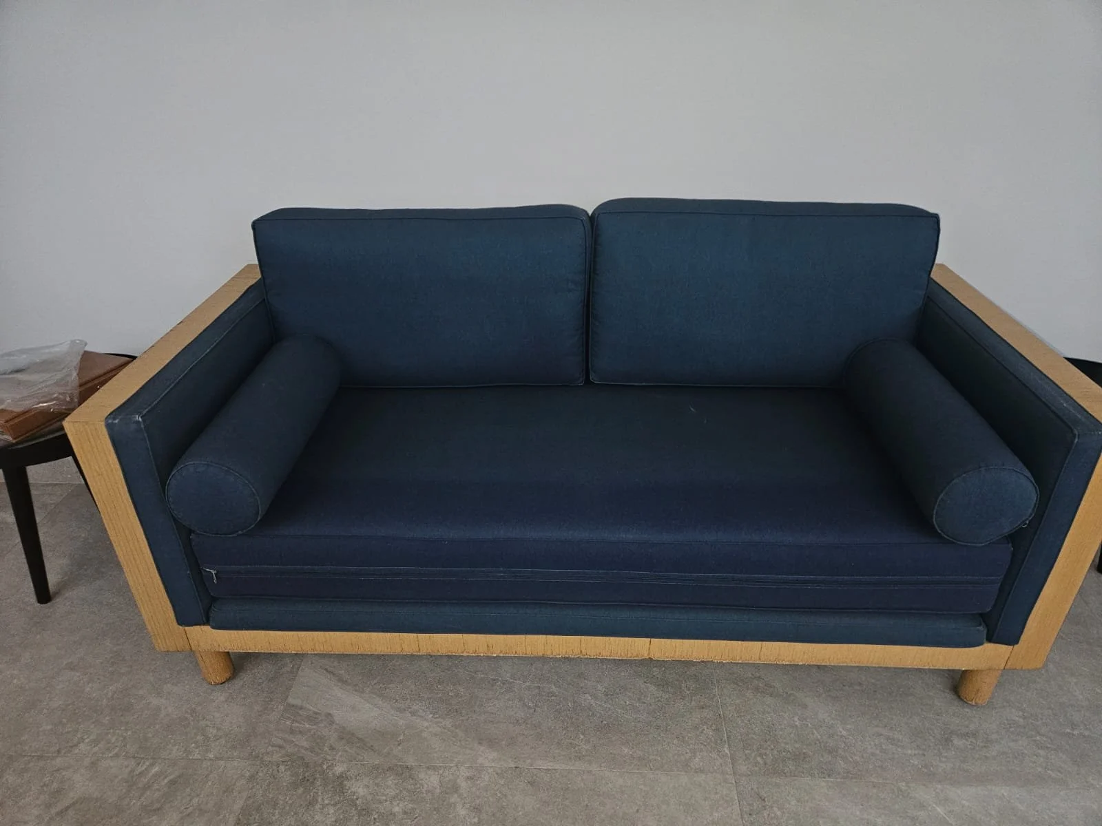 Sofá de madeira com almofadas azuis escuro e dois almofadões cilíndricos também azuis, colocado sobre um piso de ladrilhos cinzas, ao lado de uma mesa pequena com objetos.