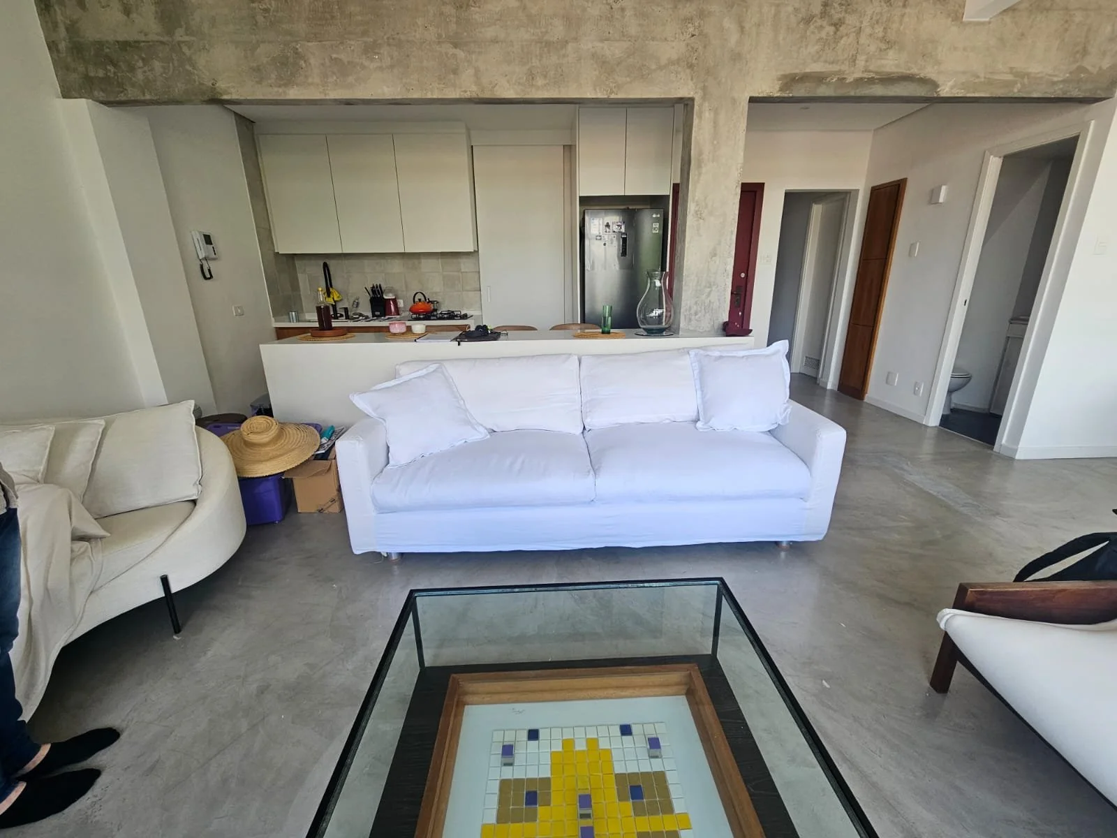 Sala de estar com sofá branco, mesa de centro de vidro e uma cozinha ao fundo com bancada e utensílios.