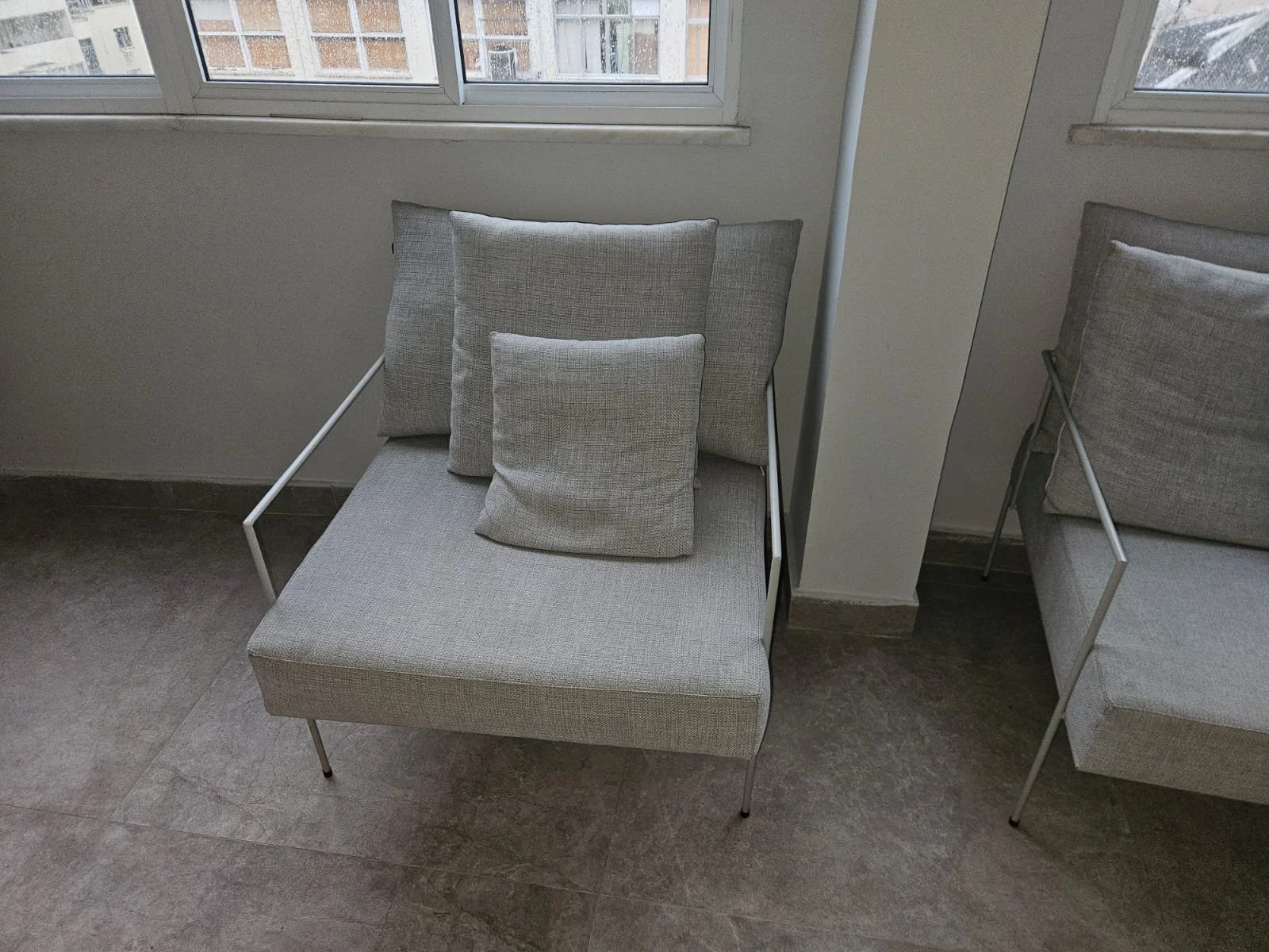 Cadeira moderna com estrutura metálica cinza e almofadas cinza clara, localizada ao lado de uma parede branca e perto de uma janela com vidro.