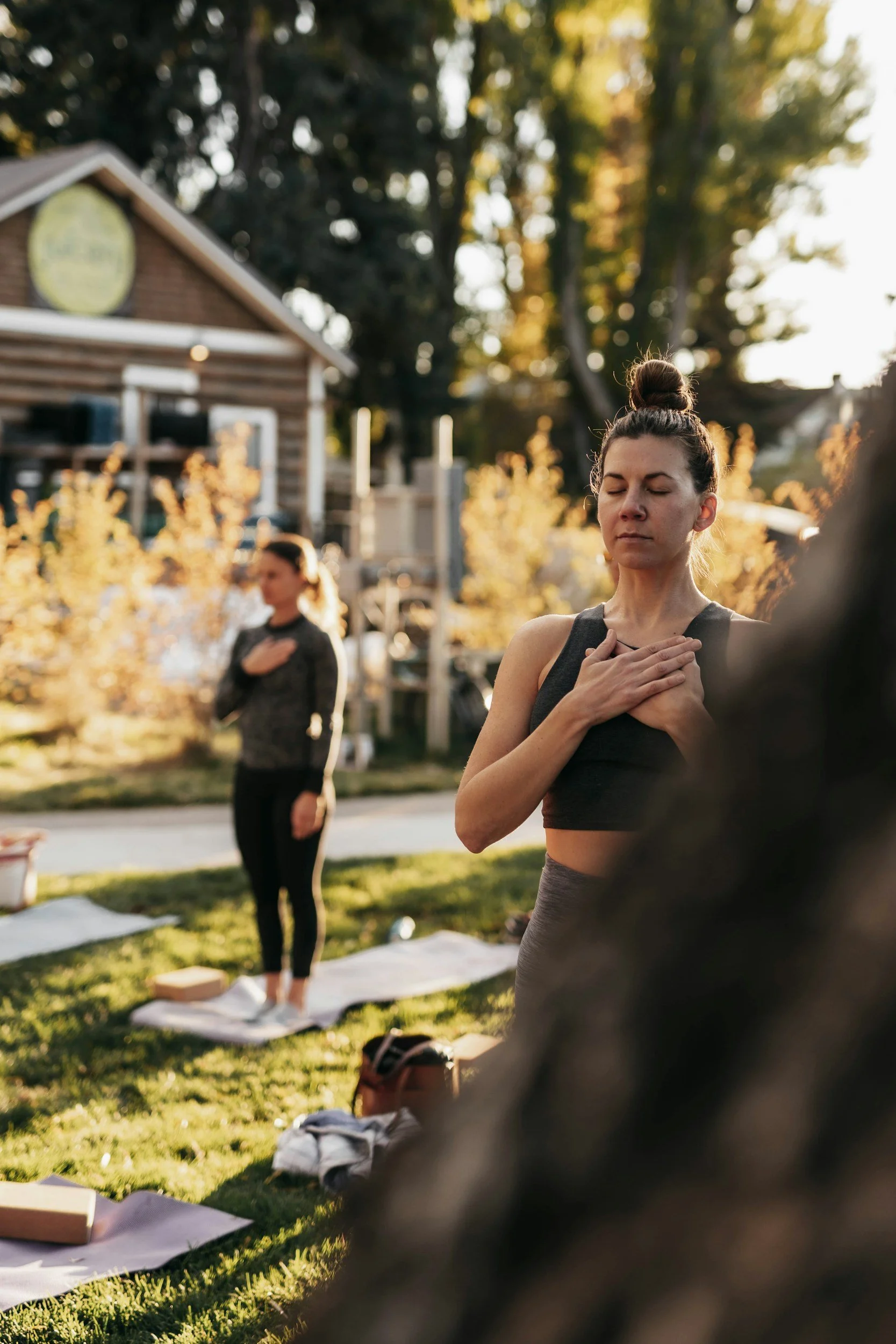 Two women practicing yoga outdoors in a garden during sunset, with open eyes and hands on their hearts, standing on yoga mats.