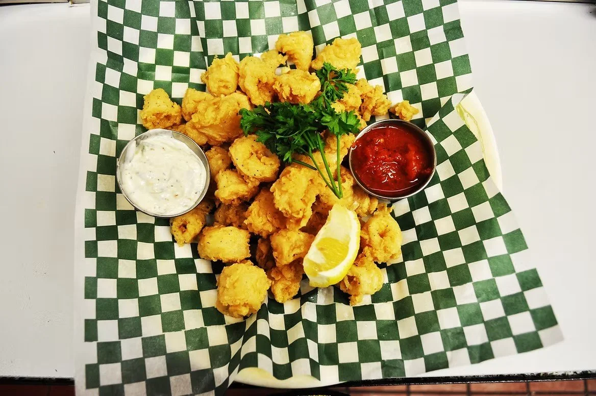 Battered fried fish pieces served on checkered paper with tartar sauce, ketchup, a lemon wedge, and parsley garnish.
