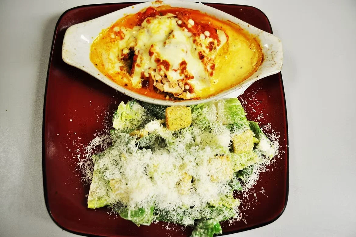 Lettuce salad topped with shredded cheese and croutons, served with baked casserole dish containing cheese, tomato sauce, and possibly chicken or meat, on a dark red square plate