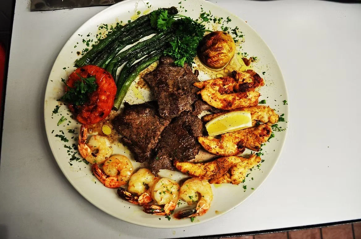 Plate of grilled shrimp, grilled chicken, grilled beef, roasted tomato, asparagus, and a lemon wedge garnished with parsley.