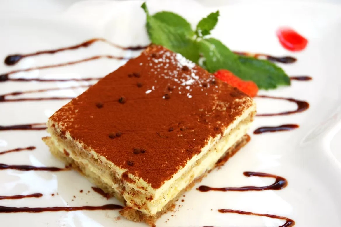 Slice of tiramisu dessert with cocoa powder, garnished with mint leaves, strawberry, and drizzled chocolate sauce on a white plate.