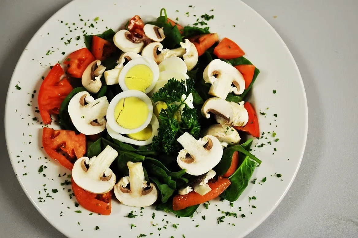 A fresh vegetable salad with sliced mushrooms, chopped tomatoes, sliced boiled eggs, spinach leaves, garnished with parsley and chopped herbs.