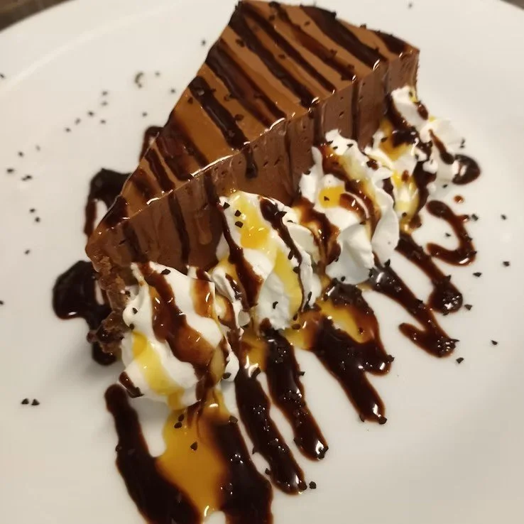 A slice of chocolate cake with whipped cream, drizzled with chocolate and caramel sauces on a white plate.