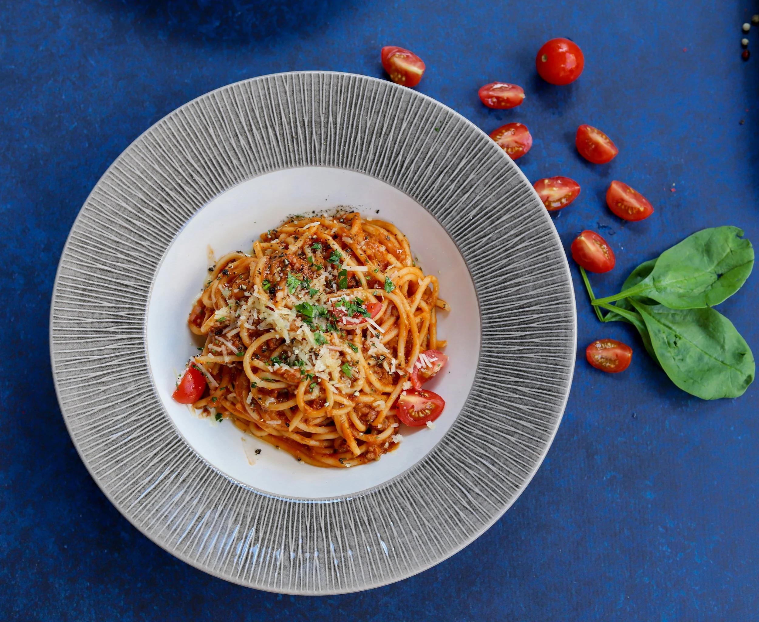 Plate of spaghetti with tomato sauce, cherry tomatoes, grated cheese, and herbs, on a decorative gray plate on a blue surface, with cherry tomato slices and basil leaves nearby.