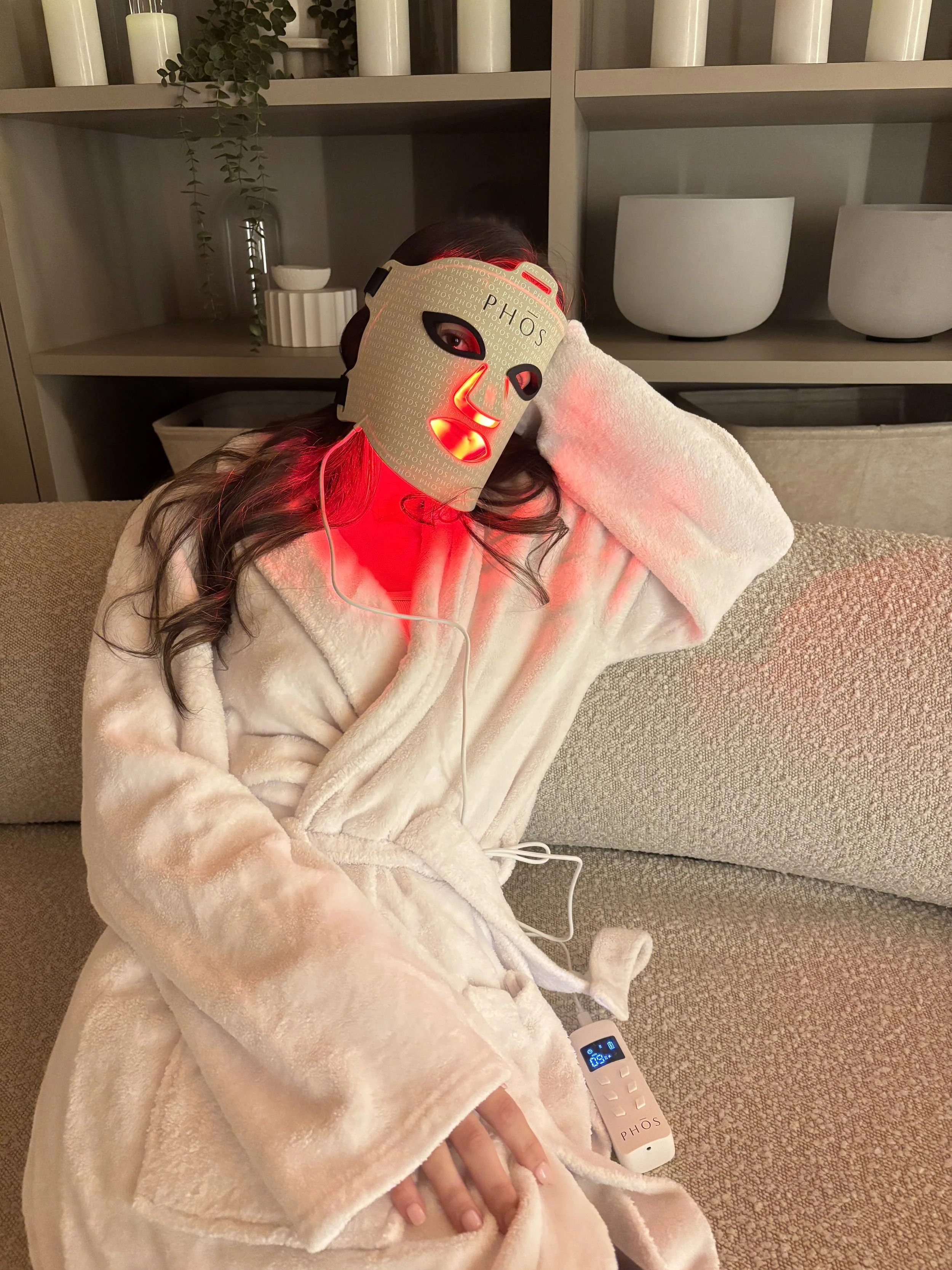 Person sitting on a beige couch, wearing a white plush robe, with a mask labeled 'PHOS' over their face, emitting red light. The person appears to be relaxing with one hand behind their head, and a device with a digital display is resting on their lap. The background includes a shelf with potted plants, candles, and decorative bowls.