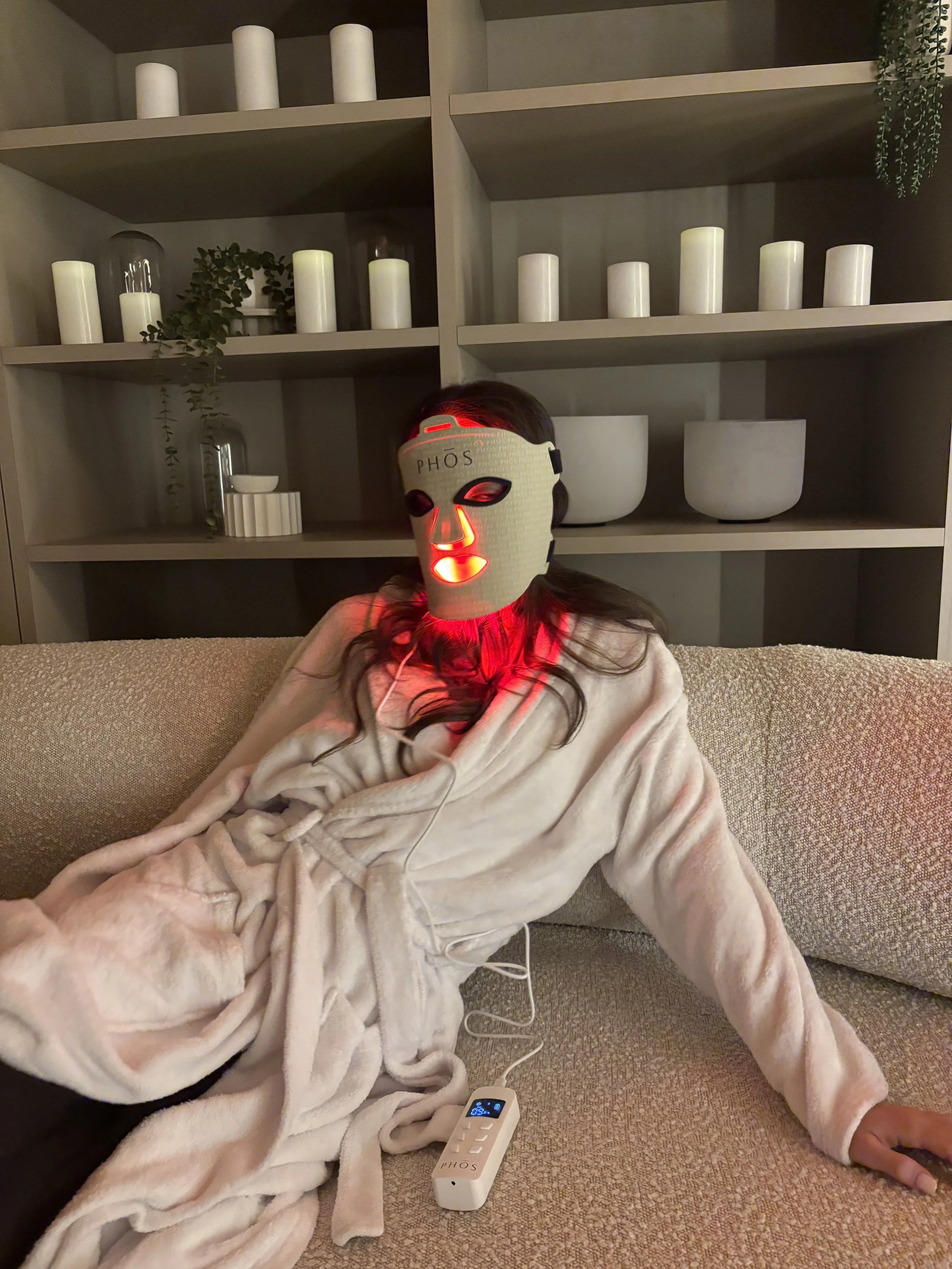 A woman lounging on a beige couch wearing a facial mask and a robe, connected to a red LED light therapy device. The background features shelves with white candles and decorative items.