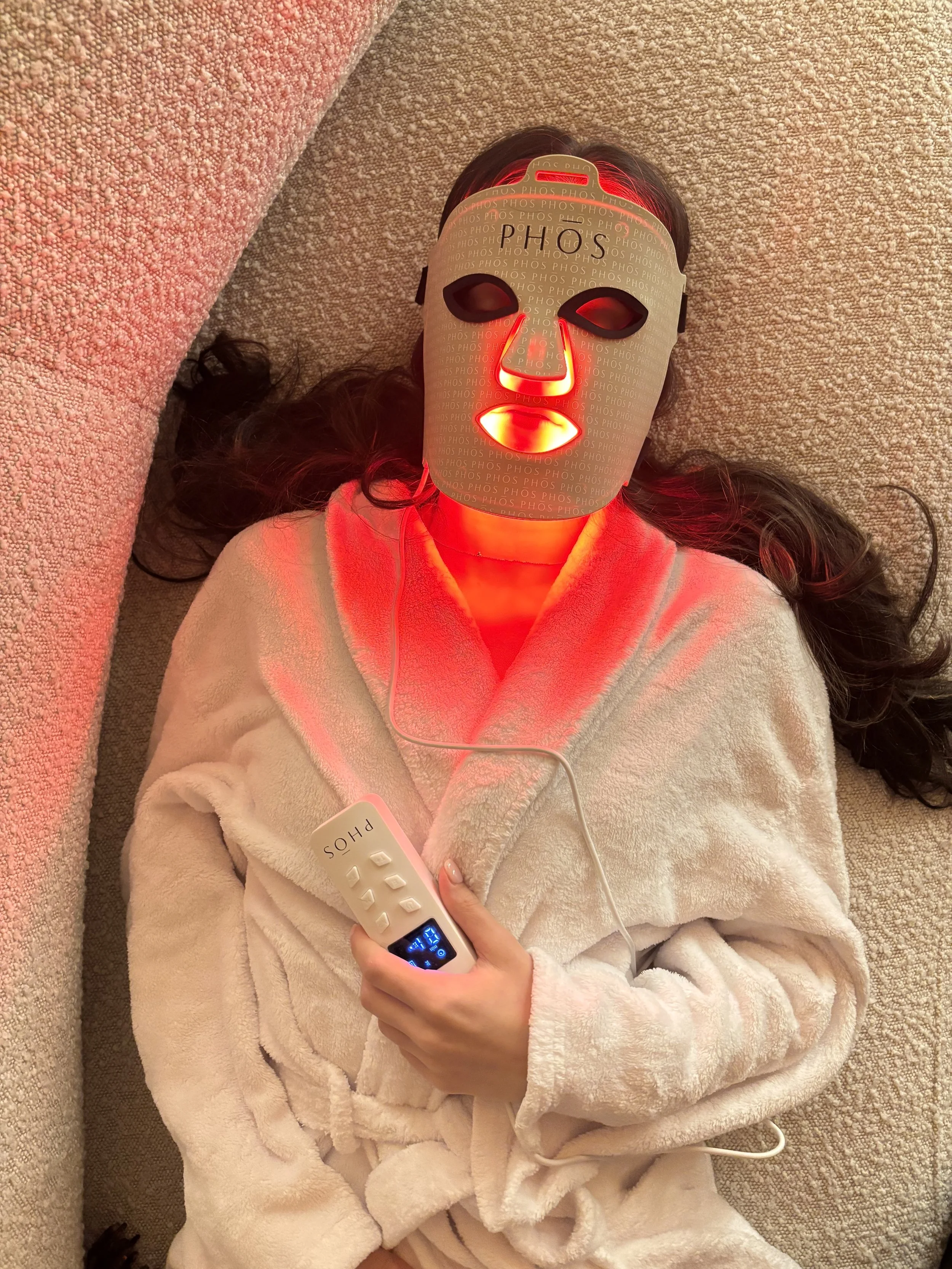 Person lying on the carpet wearing a white bathrobe with a facial mask that glows red, holding a remote control labeled 'PHO'S,