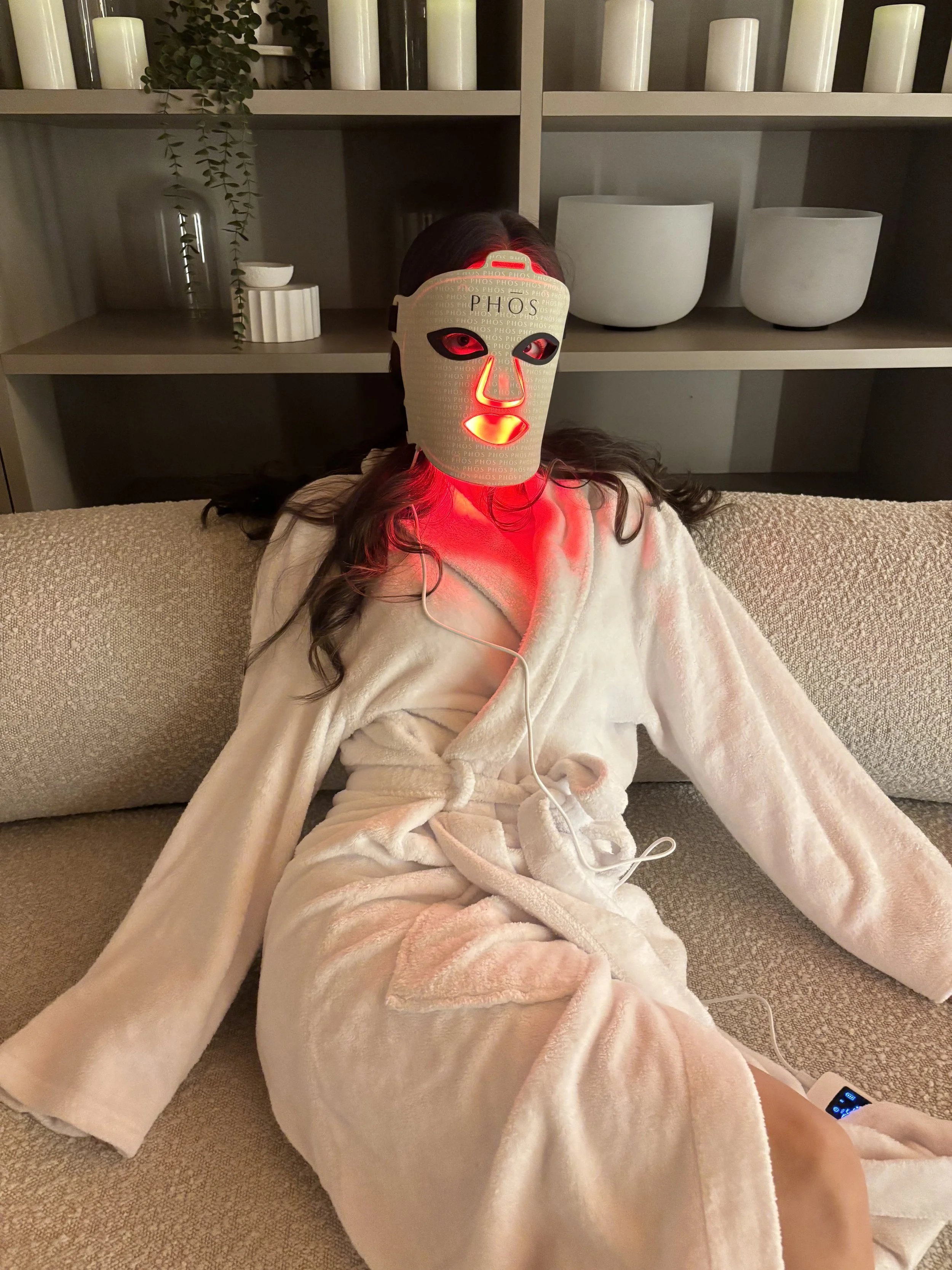 A person in a cream-colored bathrobe sitting on a beige couch with a mask of a woman's face labeled 'PHOS' over their real face. The mask has glowing red LEDs around the eyes, nose, and lips. The person is listening to music with earbuds and a device at their side.