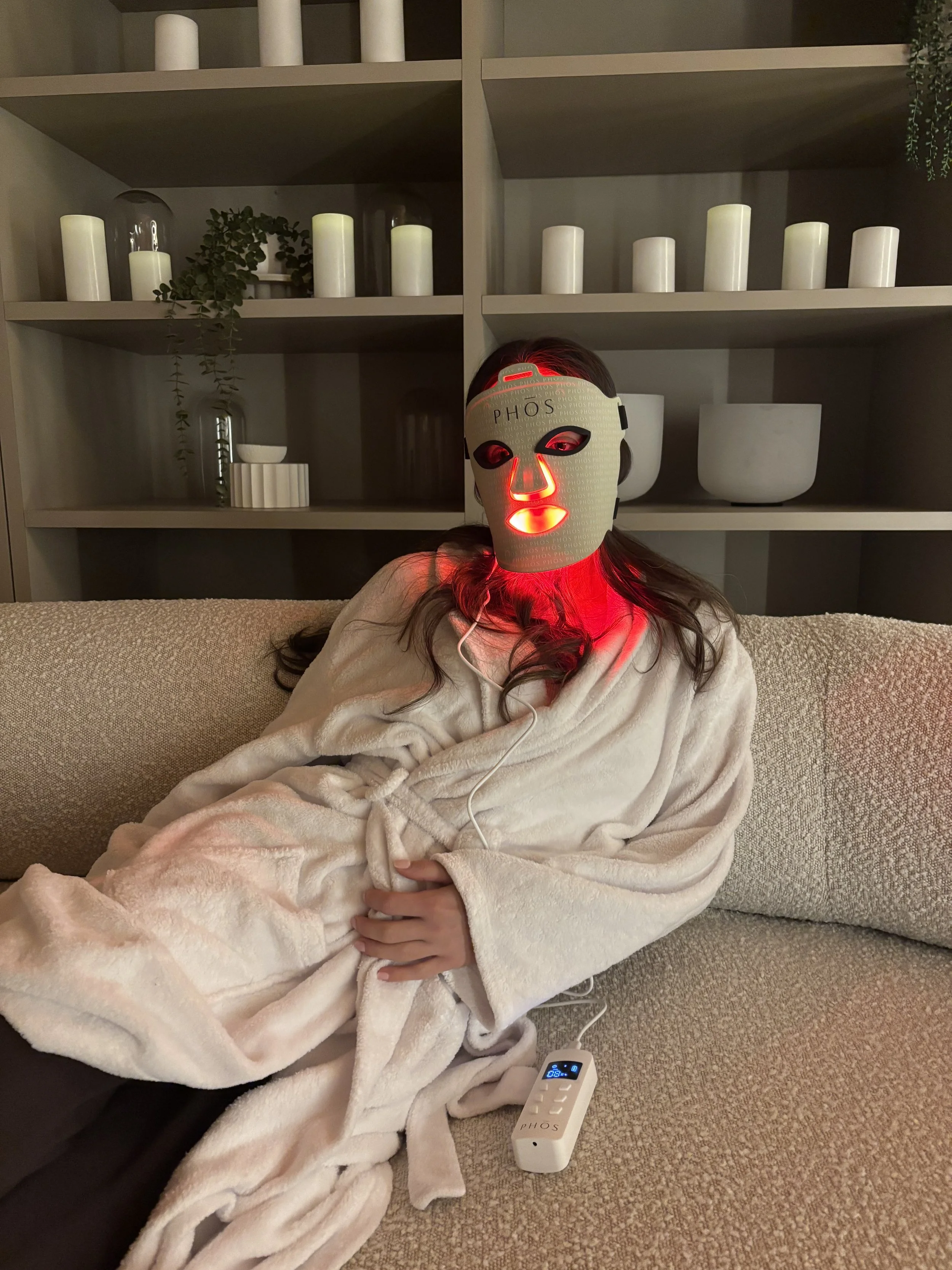 Person sitting on a sofa wearing a face-mask that is emitting red light, with a remote control and wearing a white bathrobe.