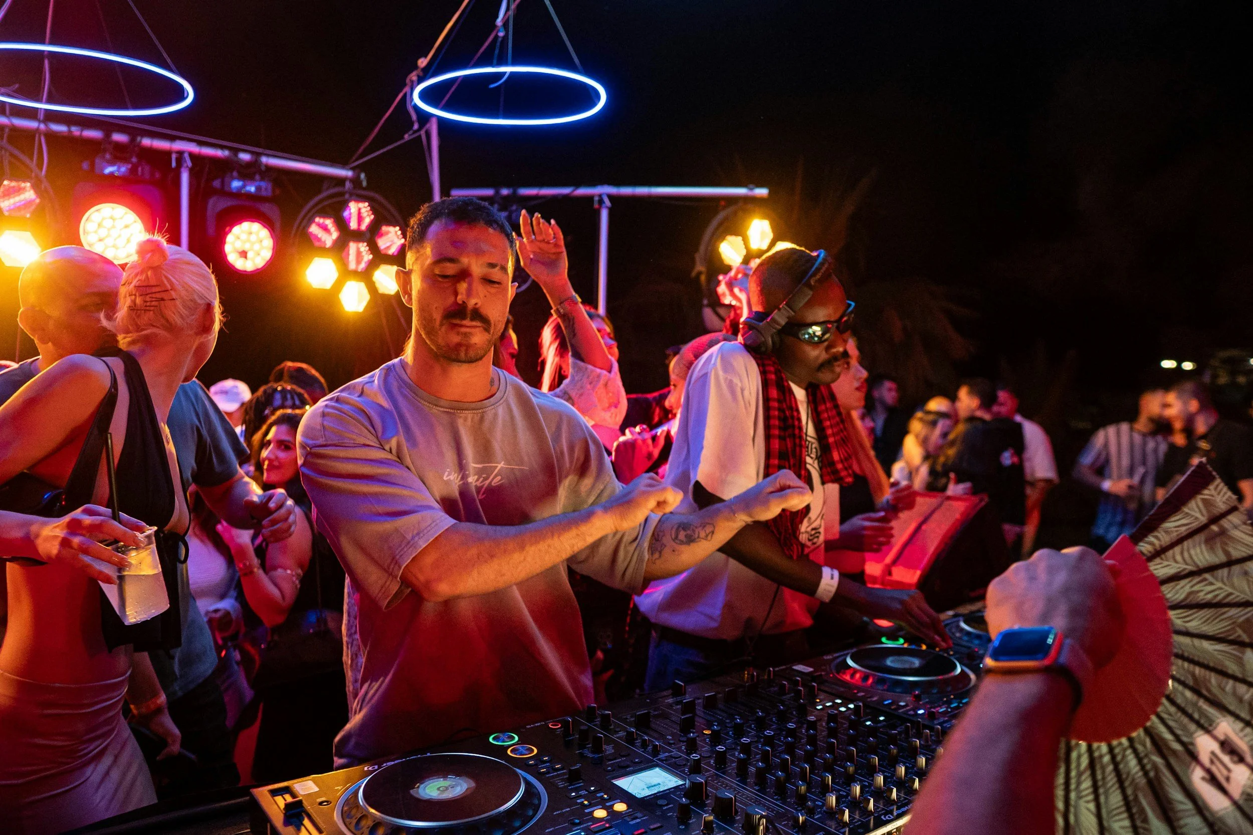 People dancing and a DJ mixing music at an outdoor nighttime party with colorful lights.