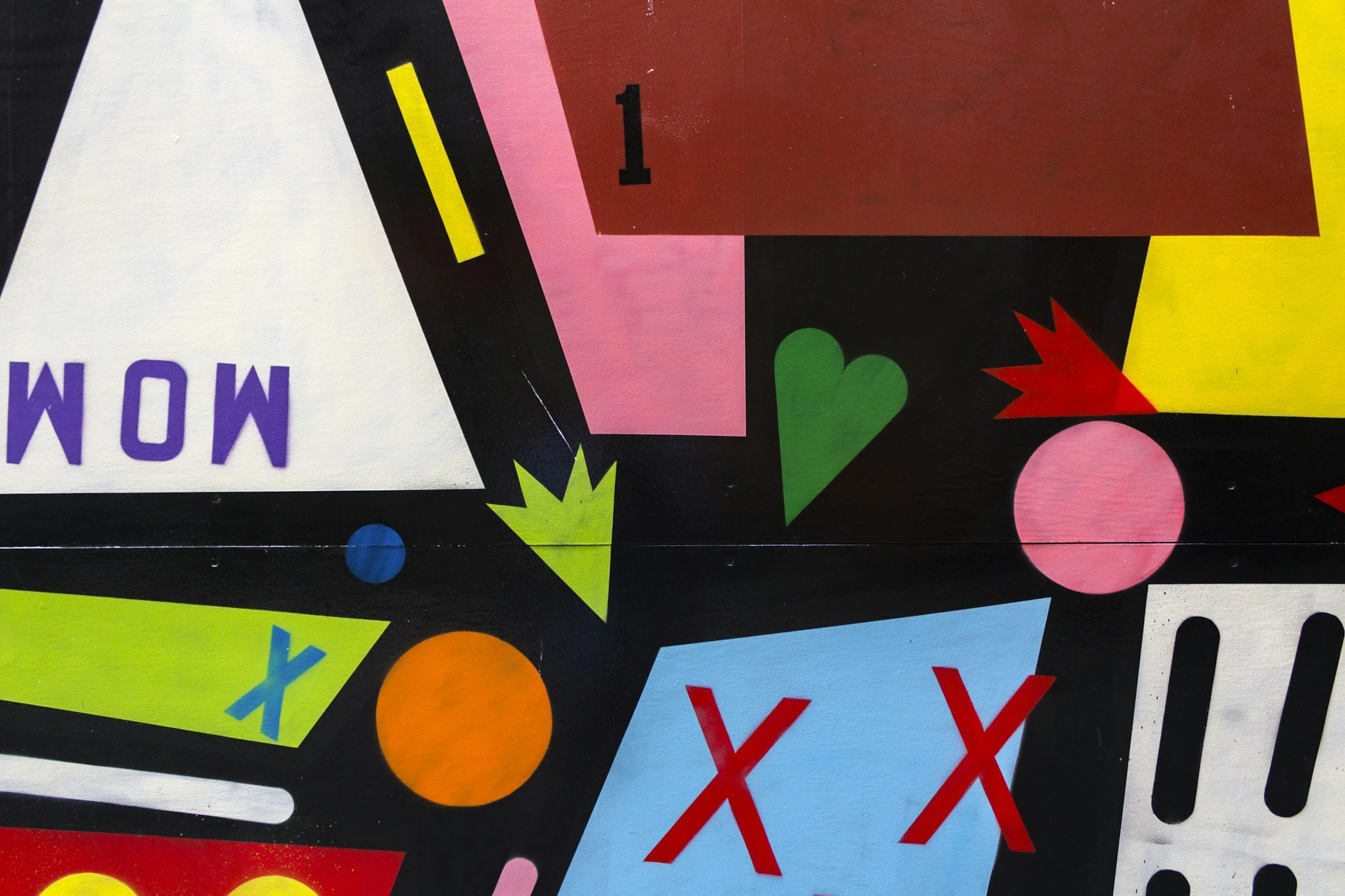 Close-up of colorful, abstract geometric shapes on a black background, including a pink triangle, yellow starburst, green heart, orange circle, and red X symbols.