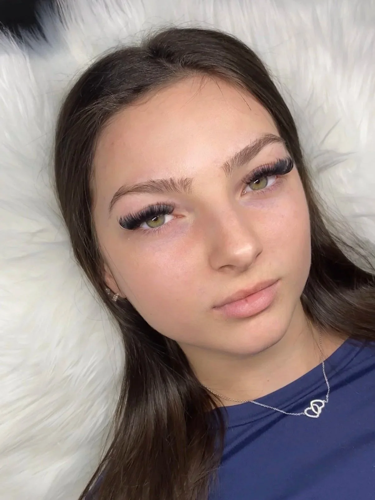 A young woman with long brown hair, fair skin, and green eyes lying on a white furry surface, wearing black eyelash extensions, small earrings, a navy blue top, and a silver necklace with a double heart pendant.