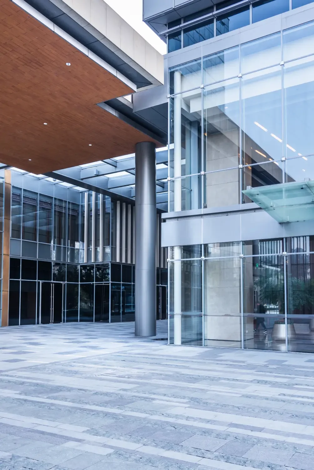 Modern building with glass walls, metal accents, and a wooden ceiling over a paved plaza.