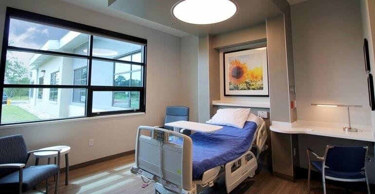 Hospital room with a bed, medical equipment, and a large window showing the outside view of a building and greenery.