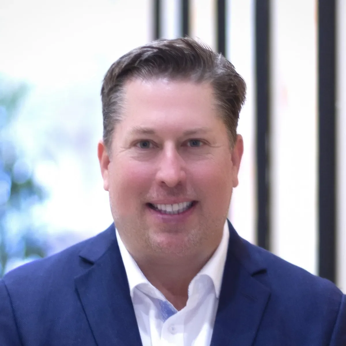 Close-up of a smiling man with neatly styled brown hair, wearing a dark blue suit jacket and a white shirt, with a blurred office background.
