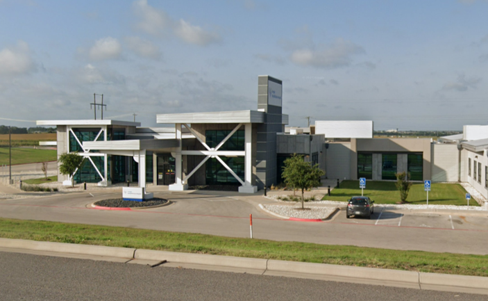 Modern building with glass windows, white exterior, and structural supports, parking lot with two handicap spaces, and a few trees, under a partly cloudy sky.