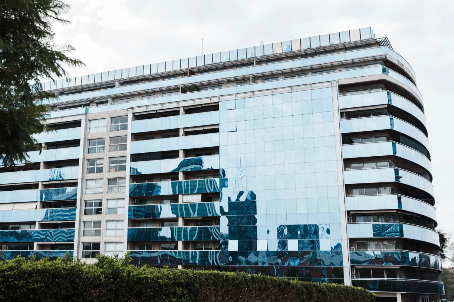 Modern multi-story building with curved balconies and a reflective glass facade, partly covered in a mural with blue and white abstract patterns, surrounded by greenery.