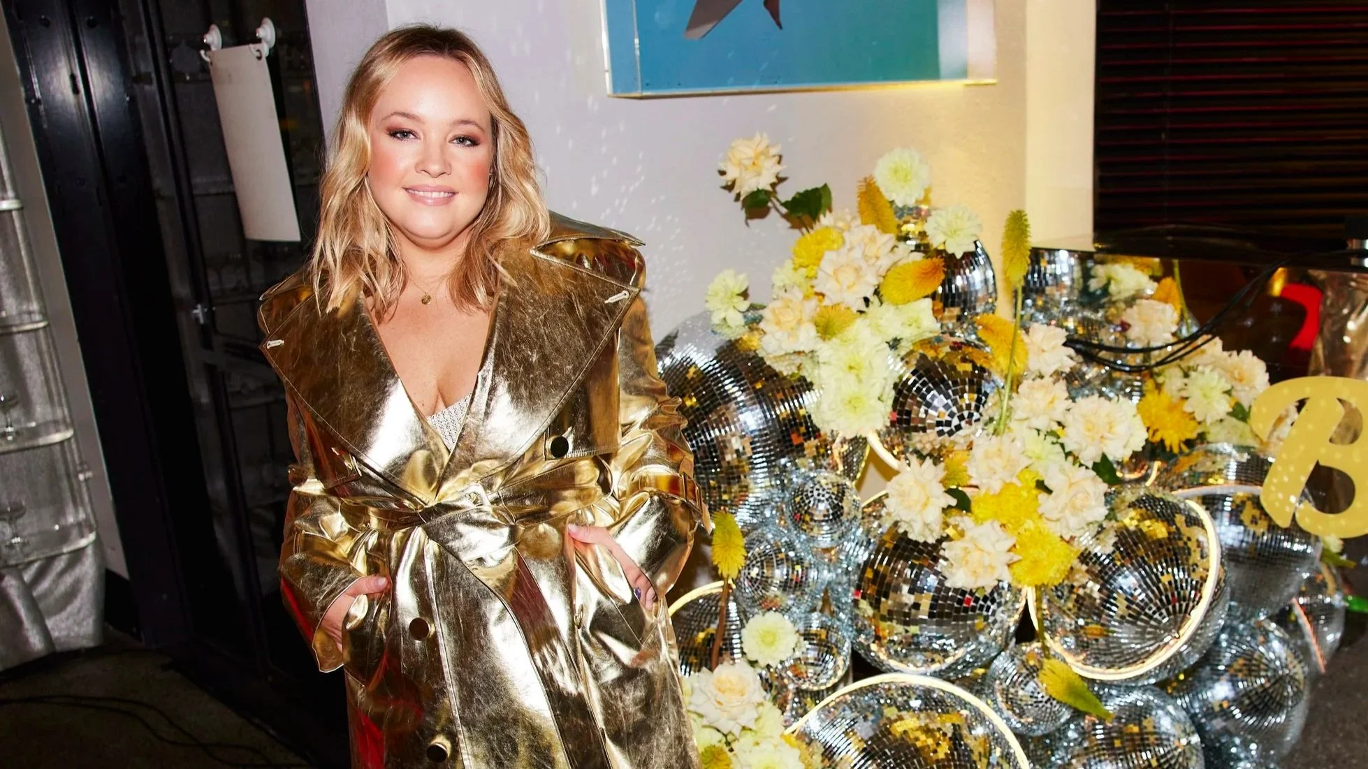 Lucille McCart, founder of Bite Size Comms, strategic communications advisor, wearing a shiny gold trench coat and standing next to a decorated table with flowers and disco balls.