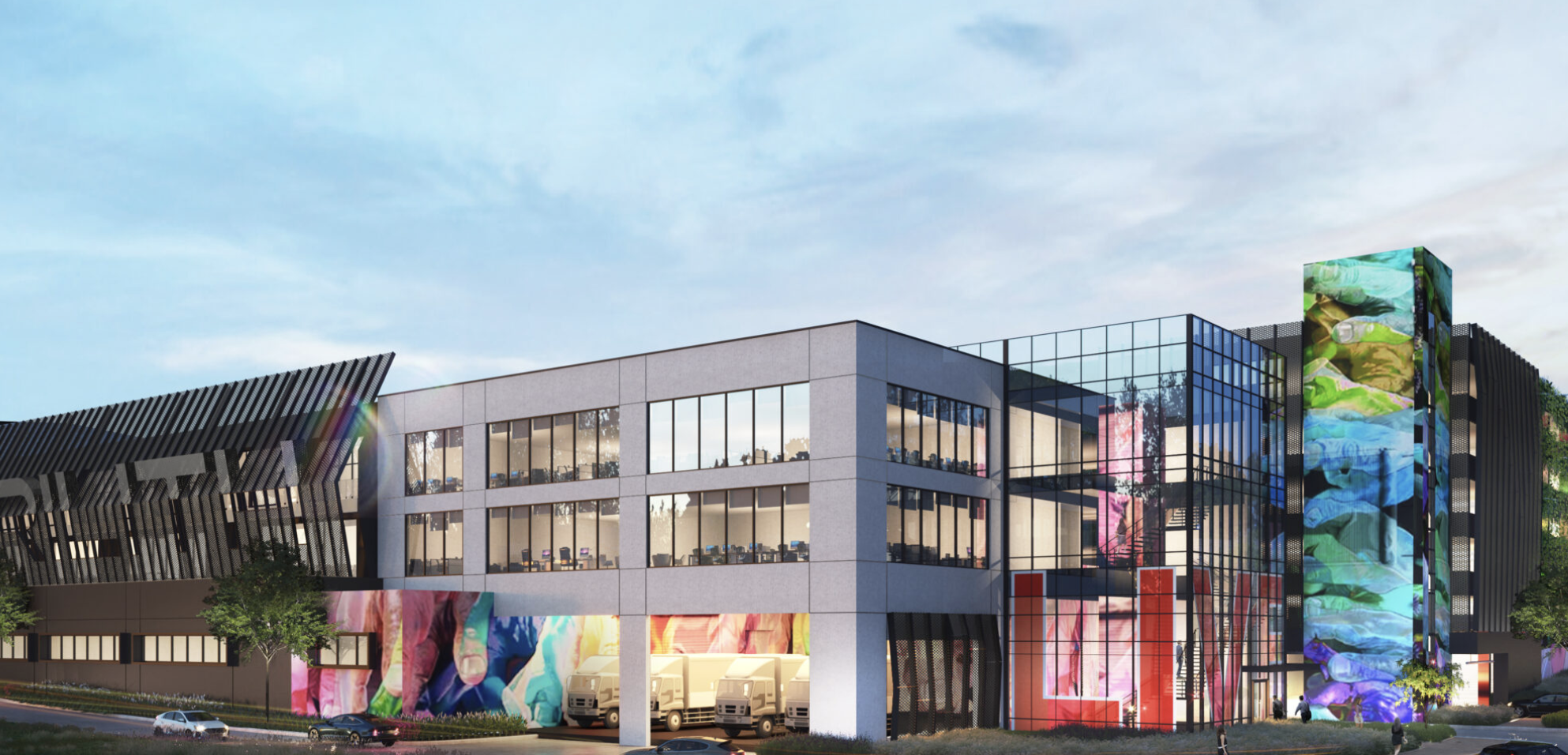 Modern multi-story building with glass and concrete exterior, large colorful digital murals, and a parking area with trucks and cars, under a partly cloudy sky.