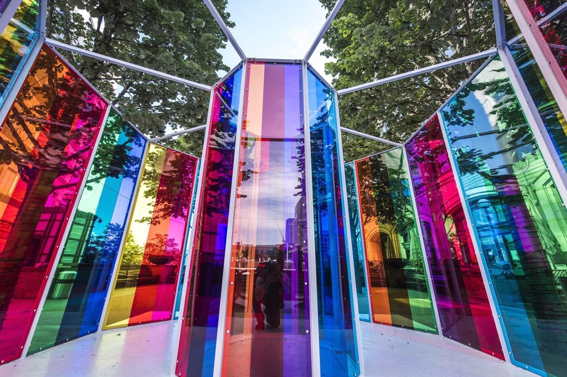 Colorful outdoor art installation with tall, vertical, rainbow-colored glass panels surrounded by trees and city buildings.