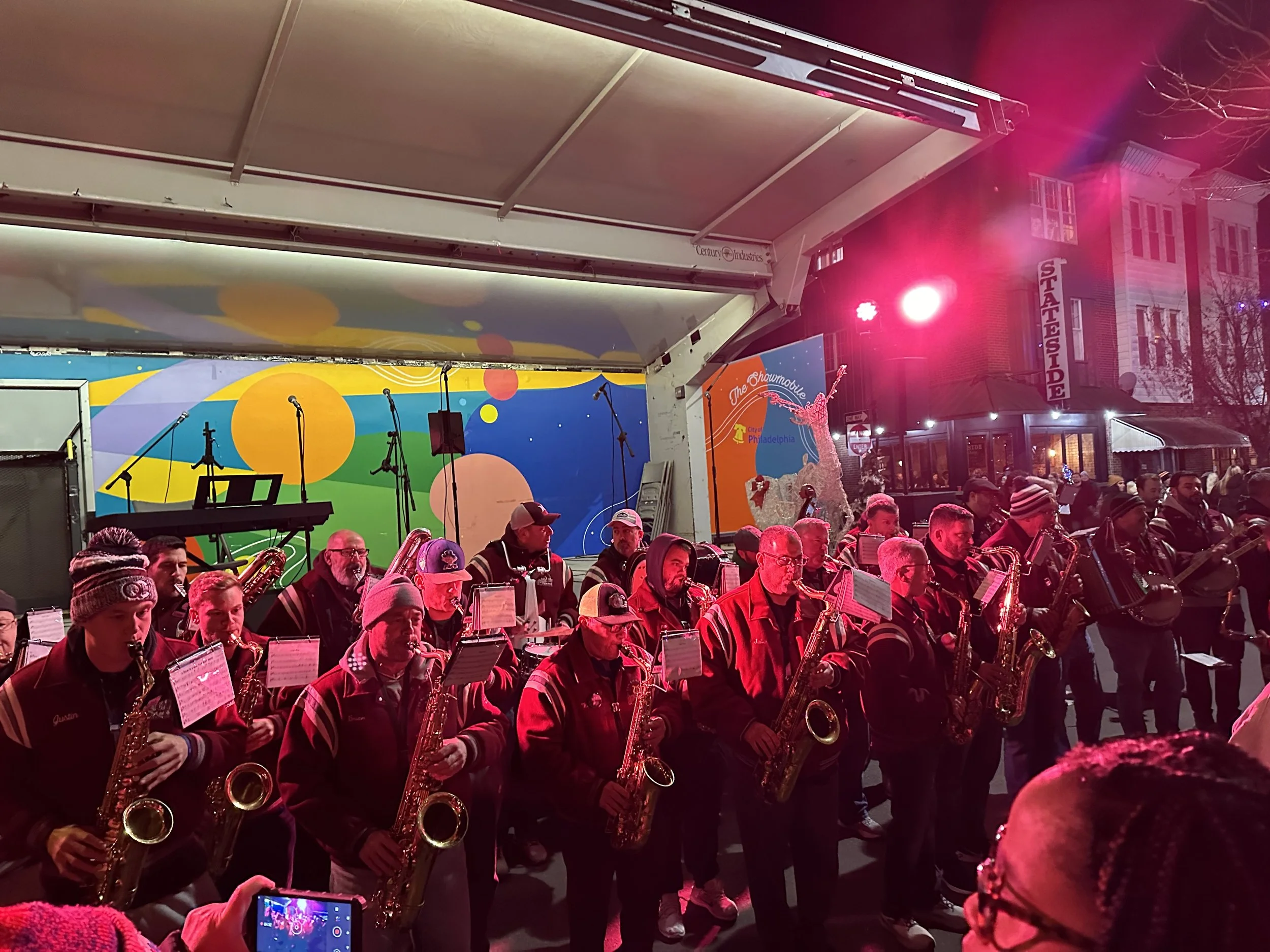A musical performance taking place outdoors at night, featuring a band with saxophonists under a decorated stage with a colorful mural backdrop. Audience members watching, some wearing winter clothing.