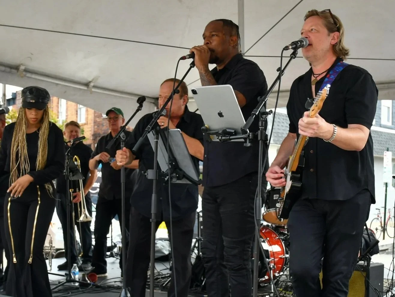 Musicians performing on stage at an outdoor event, including a woman with long dreadlocks, a man singing into a microphone, and a guitarist with glasses and a necklace.
