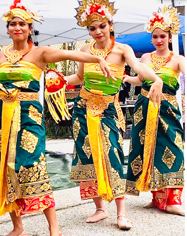 Three women performing a traditional dance, dressed in vibrant green, blue, and yellow costumes with floral crowns and jewelry, at an outdoor cultural event.