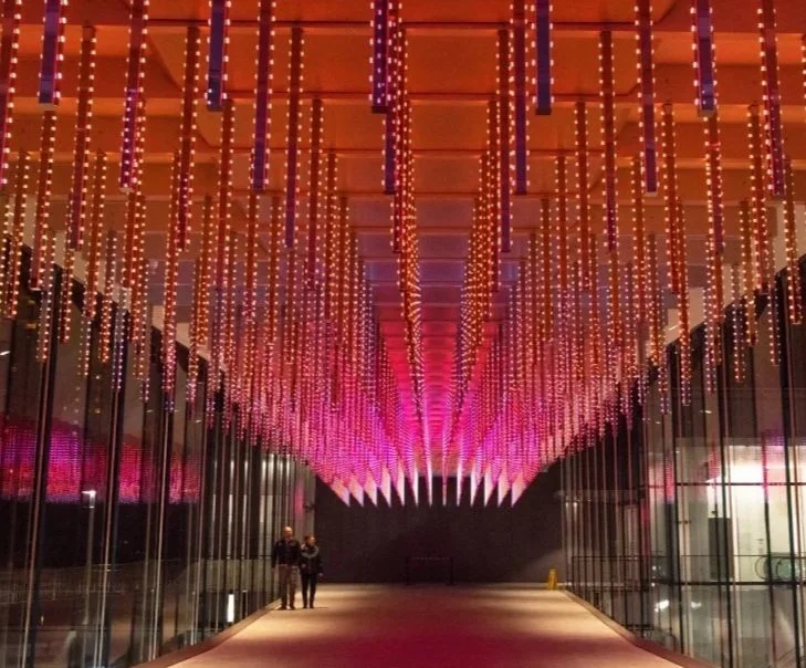 Indoor hallway with ceiling decorated with hanging lights in purple and orange hues, reflecting on glass walls and leading to a dark back wall where two people are walking.