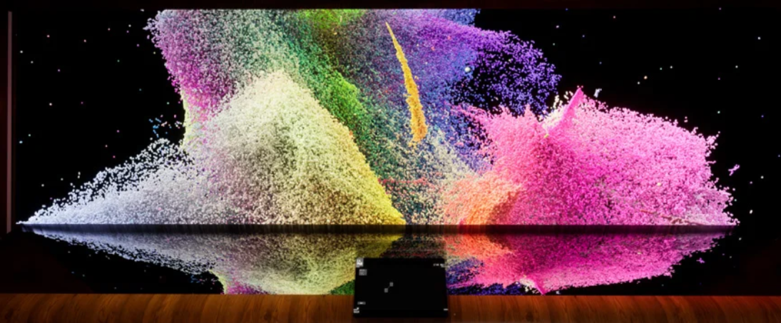 Large digital display showing colorful powder explosions in pink, purple, yellow, green, and blue, with a black background and reflections on the surface below.
