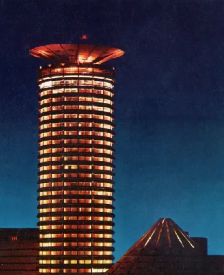 Nighttime cityscape featuring a tall, illuminated skyscraper with circular balconies and a distinctive ringed top, alongside a pyramid-shaped building with illuminated lines.