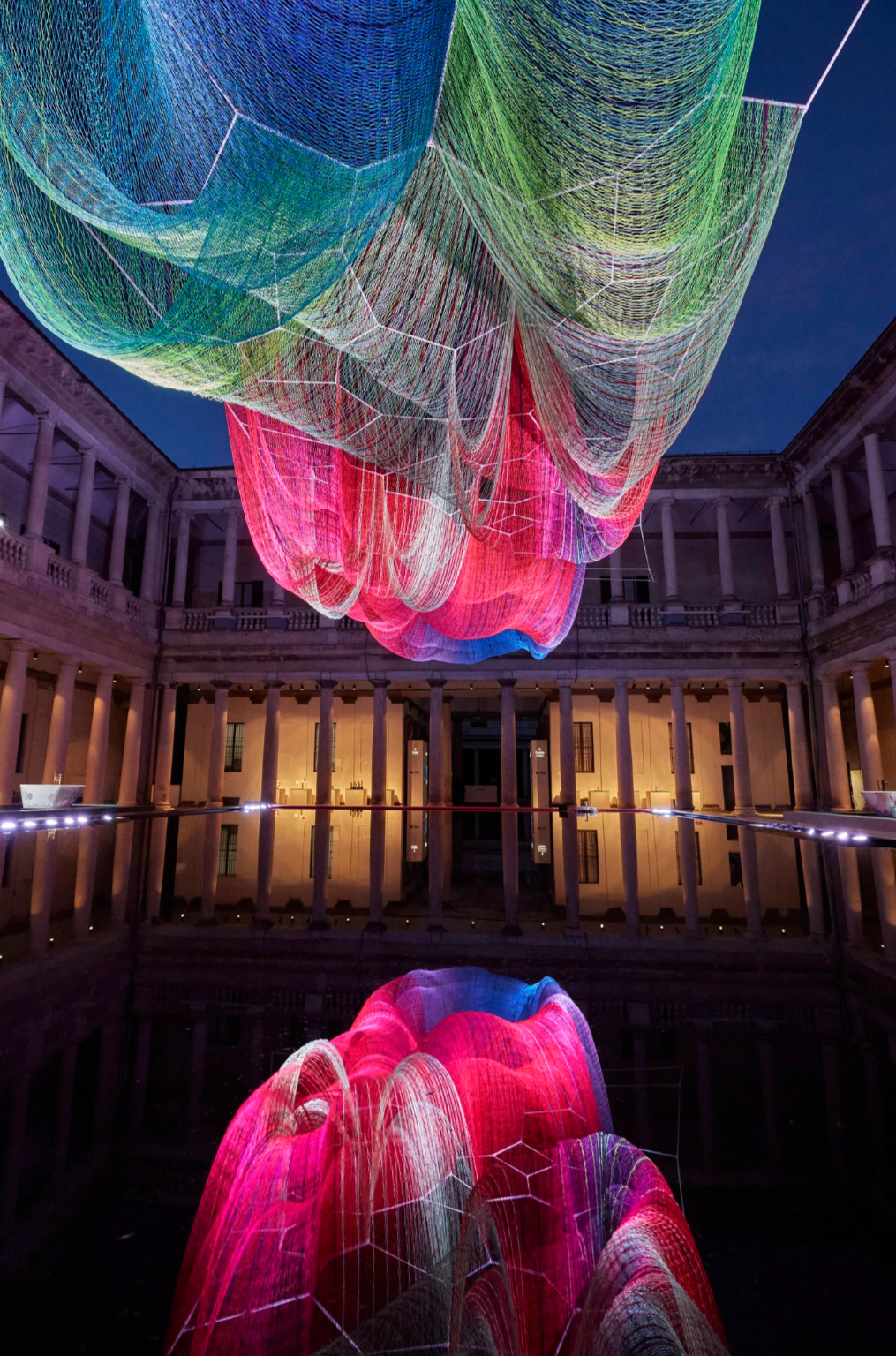 Colorful illuminated art installation hanging above a reflecting pool inside a classical building with columns, night sky in background.