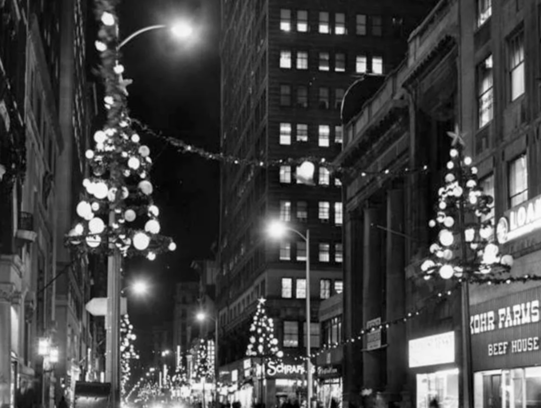 Nighttime city street decorated with Christmas trees and lights, with tall buildings and lit signs.