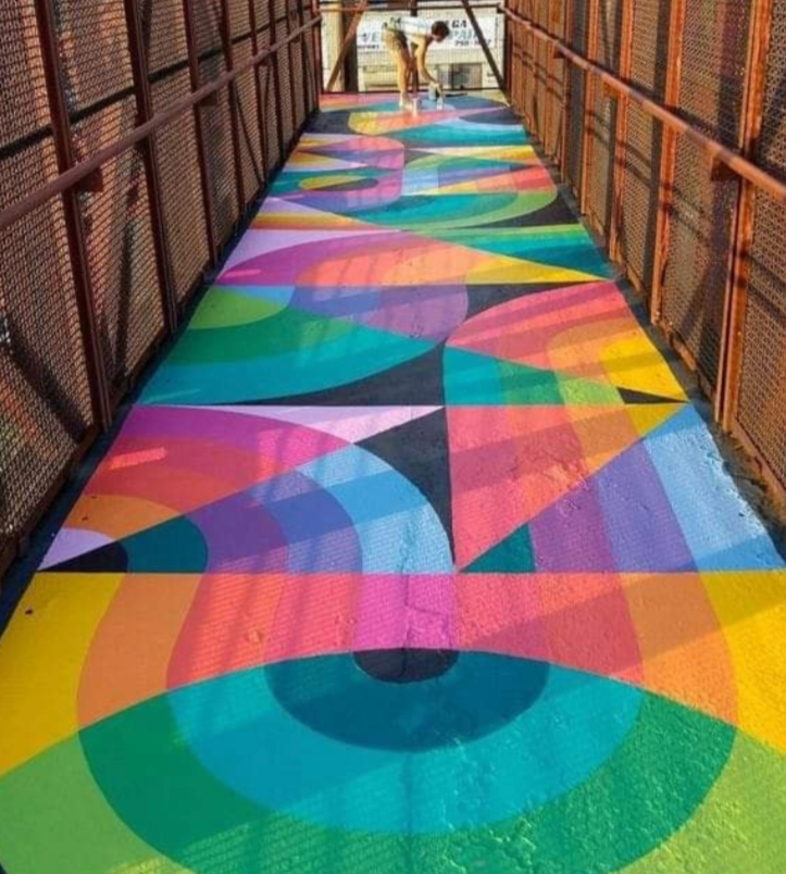 Colorful geometric mural painted on a walkway bridge with orange metal railing on sides.