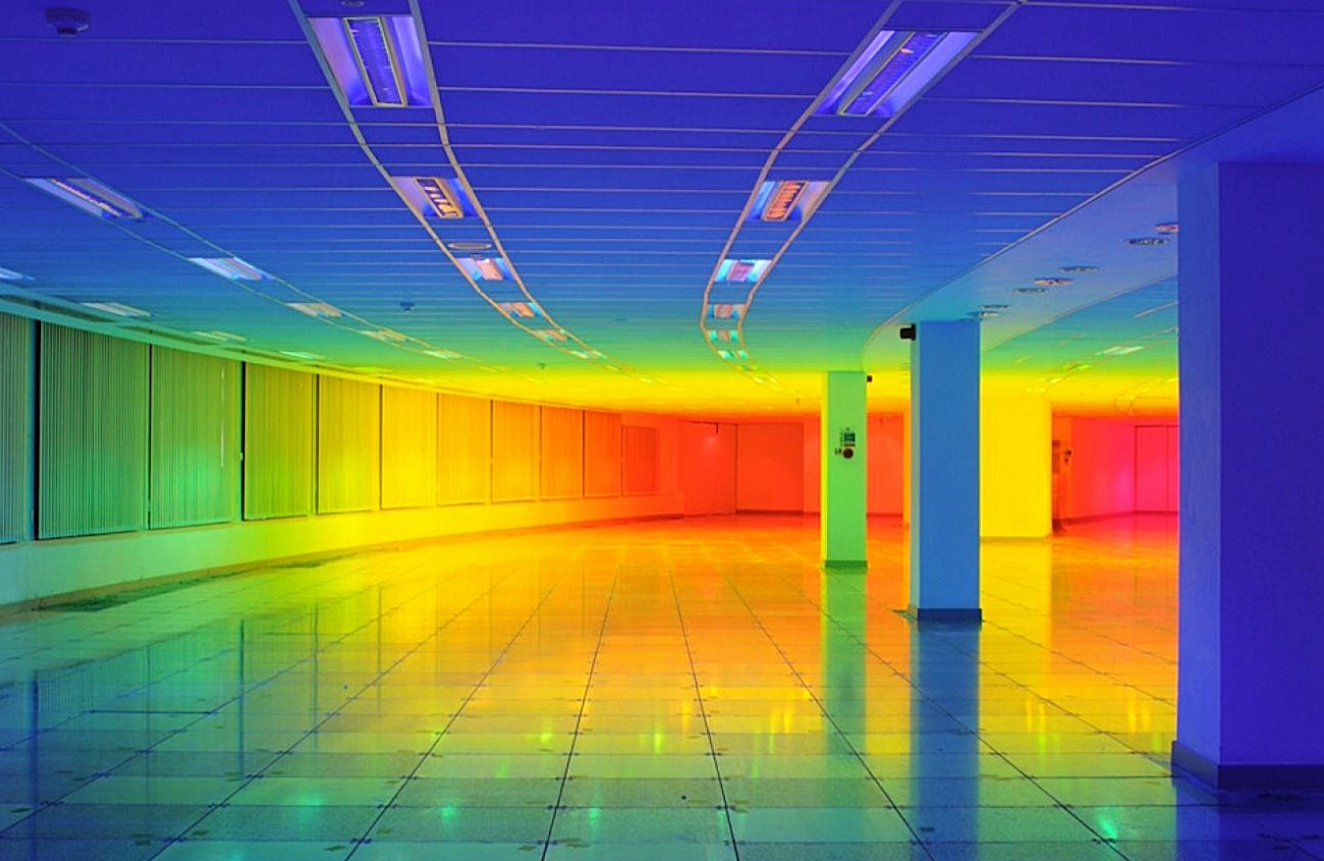 Empty indoor space with rainbow-colored lighting creating a vibrant atmosphere, with a tiled floor, white columns, and ceiling panels.