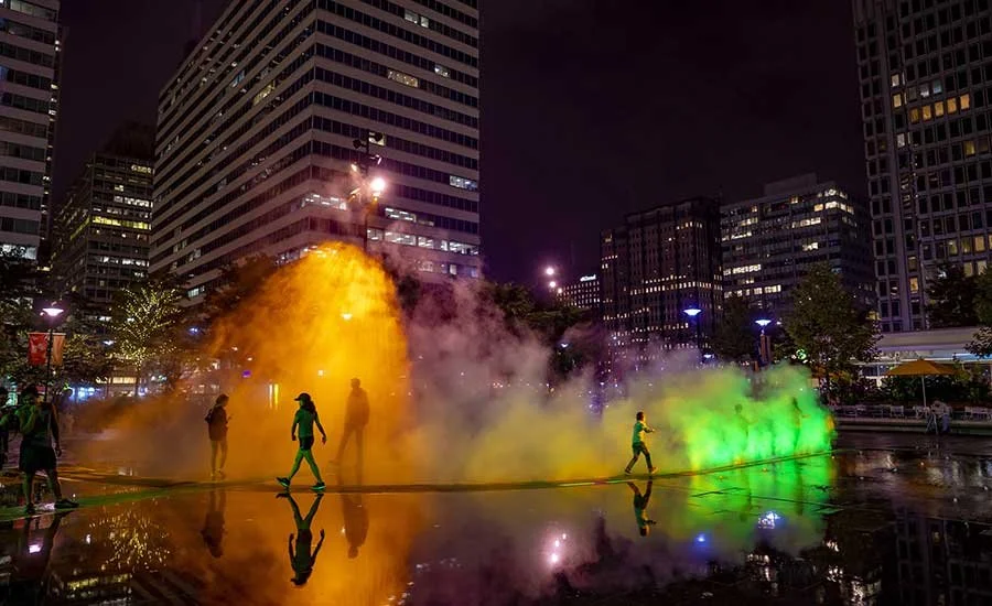 People walking through a colorful fog display in an urban park at night, with tall buildings illuminated in the background.