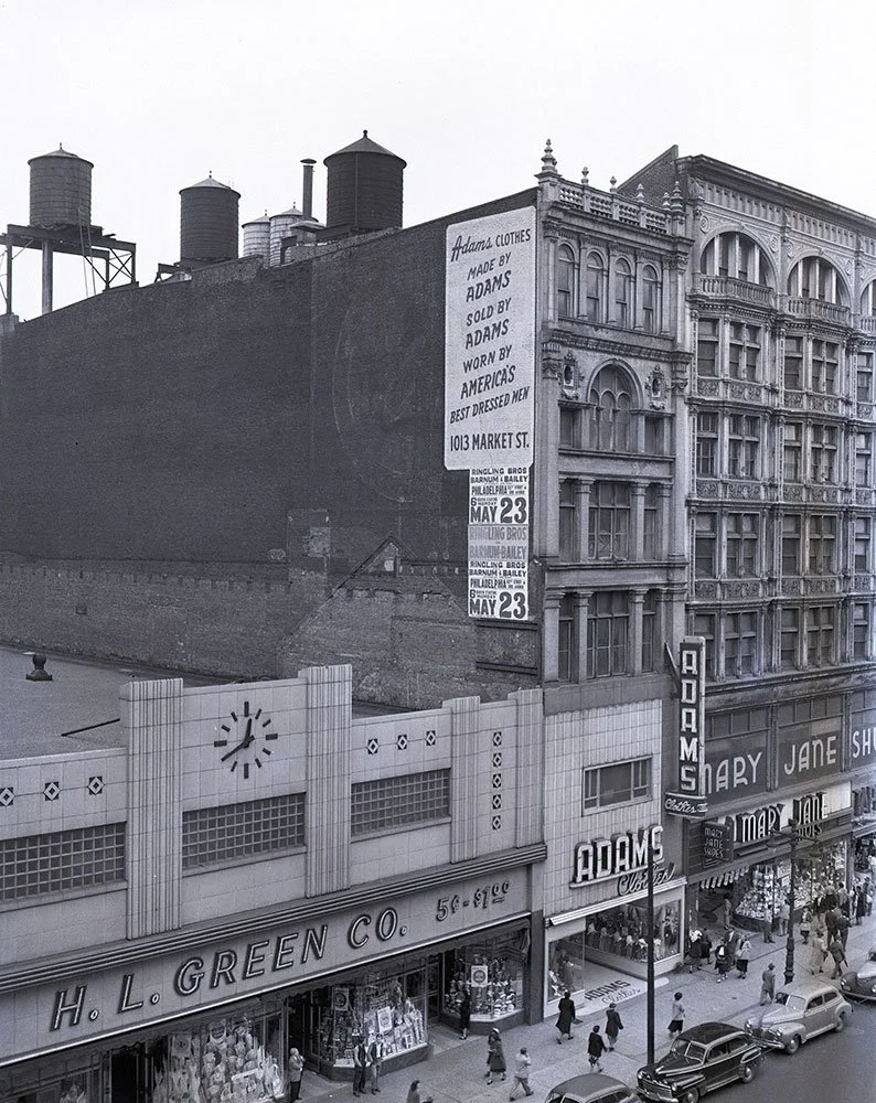 Black and white street scene showing storefronts, pedestrians, and parked cars. Prominent signage includes "H. L. GREEN CO." and "ADAMS.