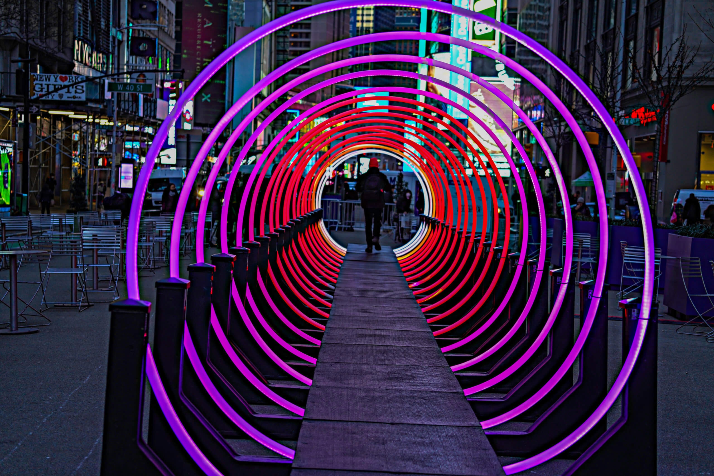 A lit circular tunnel installation on a city street at night, with a person walking through the tunnel. Neon lights in purple and red create a vibrant, colorful scene surrounded by buildings and digital billboards.