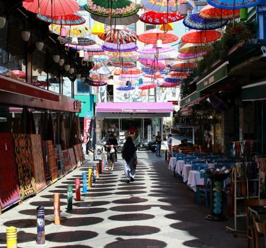 Street scene with colorful umbrellas hanging overhead, casting shadows on the walkway below, with pedestrians walking through shopping area.