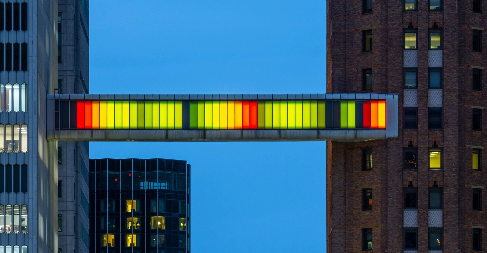 City skyline at dusk with a multicolored illuminated skywalk connecting two buildings.