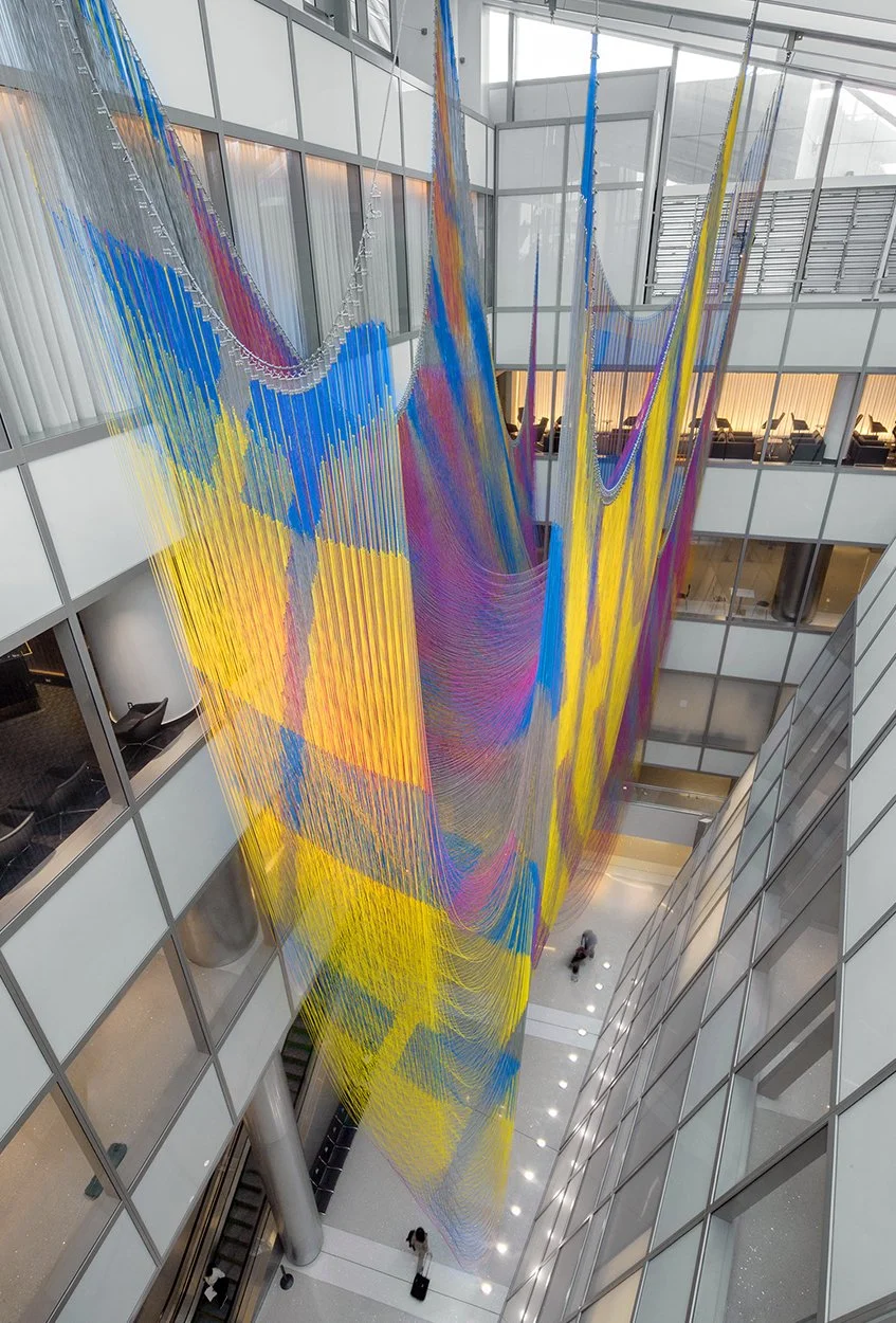 Multicolored hanging art installation inside a modern building atrium with glass walls and floors.