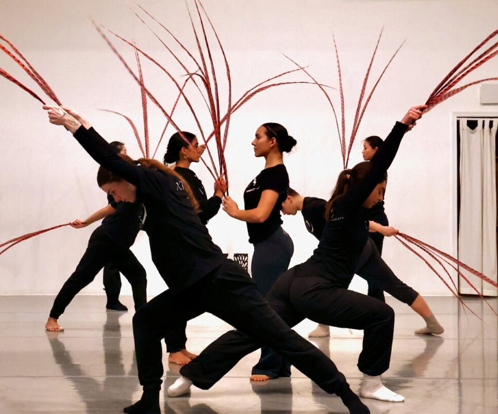 Six dancers in black outfits perform a dance with long red sticks in a white room.